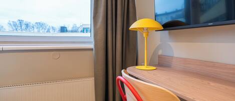A room with a wooden desk, a yellow lamp, a red chair, a window with curtains, and a view of buildings outside.