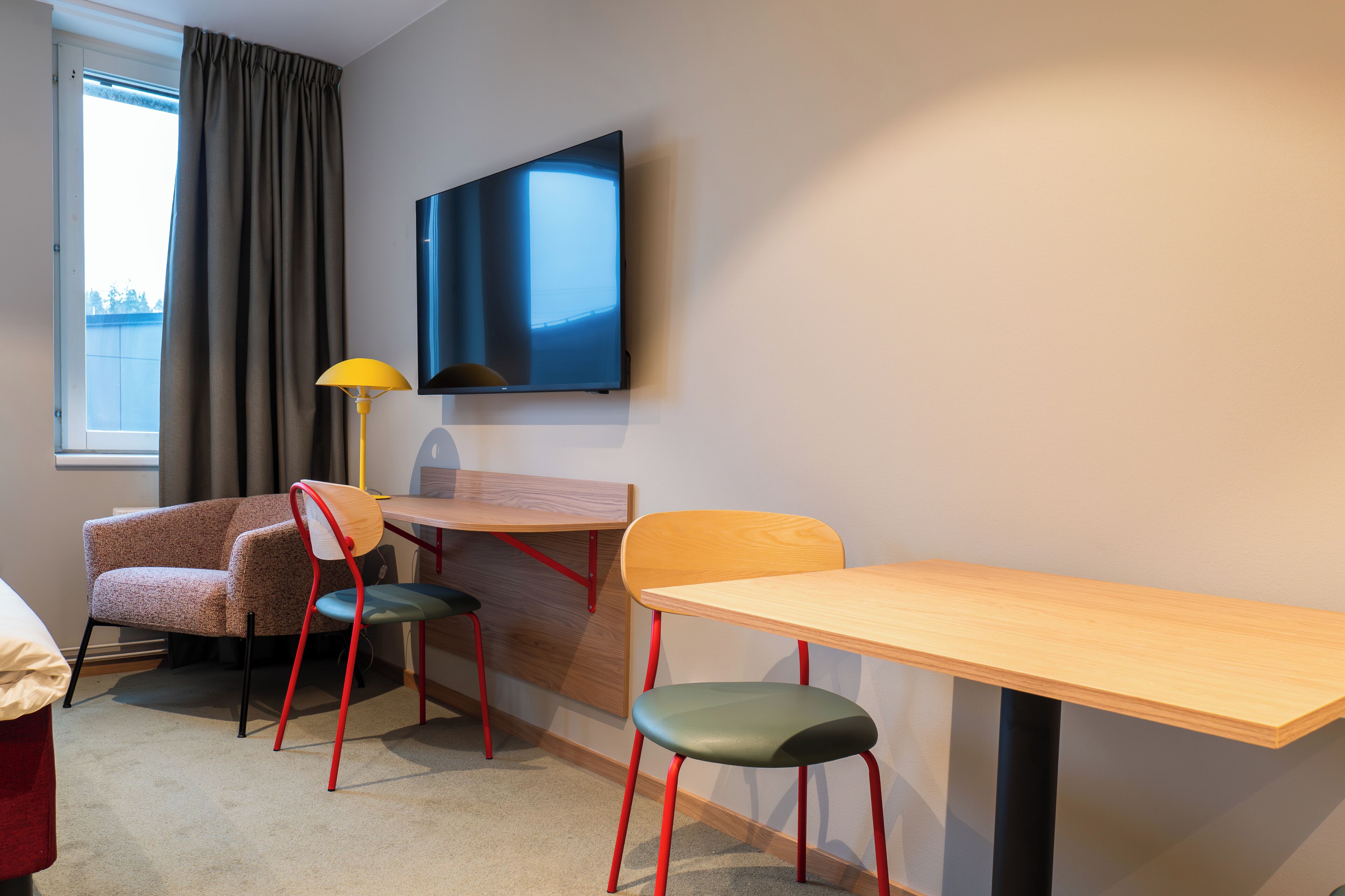 Una habitación de hotel con cama, escritorio, silla, televisor y ventana con cortinas.