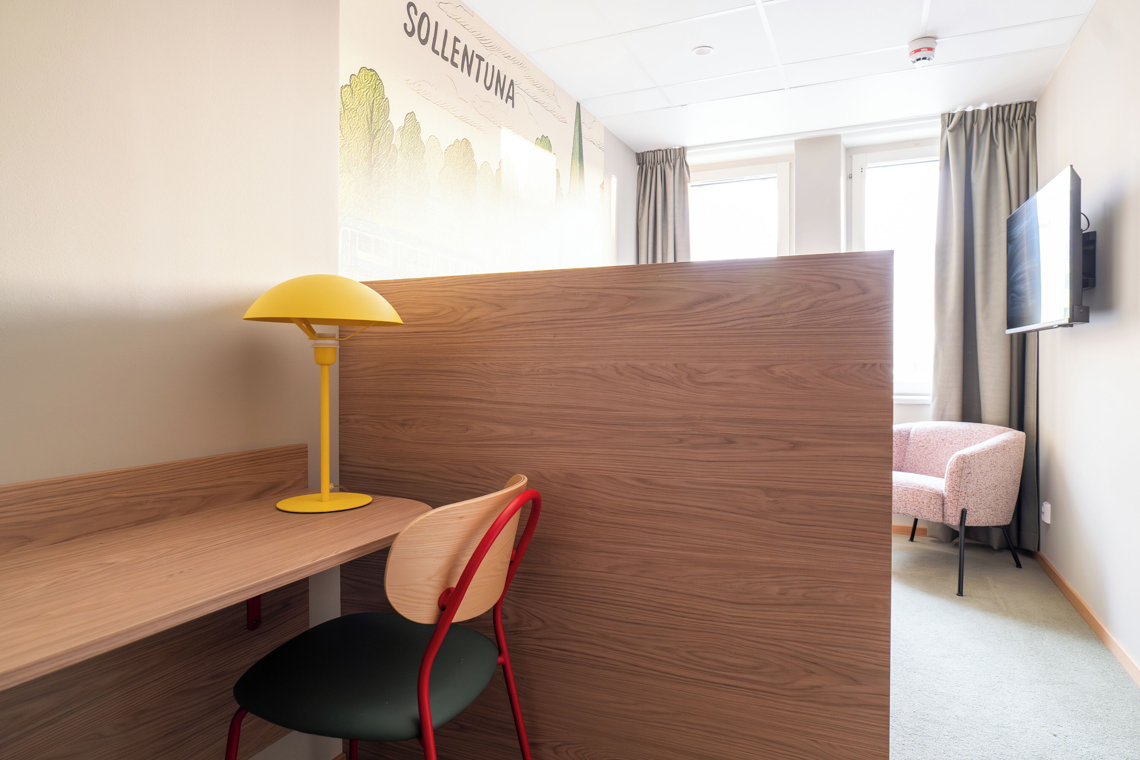A hotel room with a wooden desk, a yellow desk lamp, a red and green chair, a partition wall, a TV mounted on the wall, and a window with curtains.