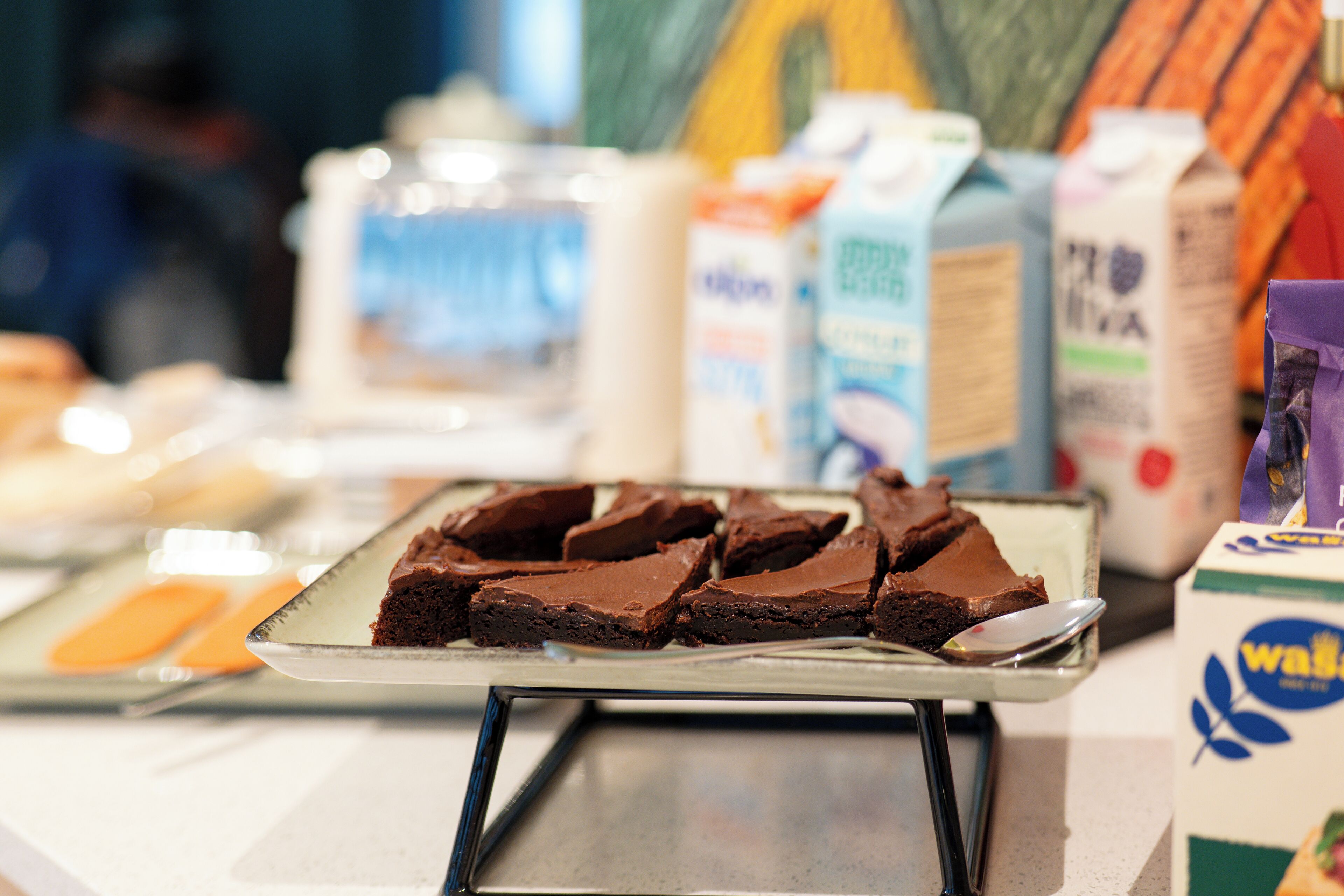 A tray of brownies on a metal stand with a spoon. In the background, there are cartons of milk and other food items.
