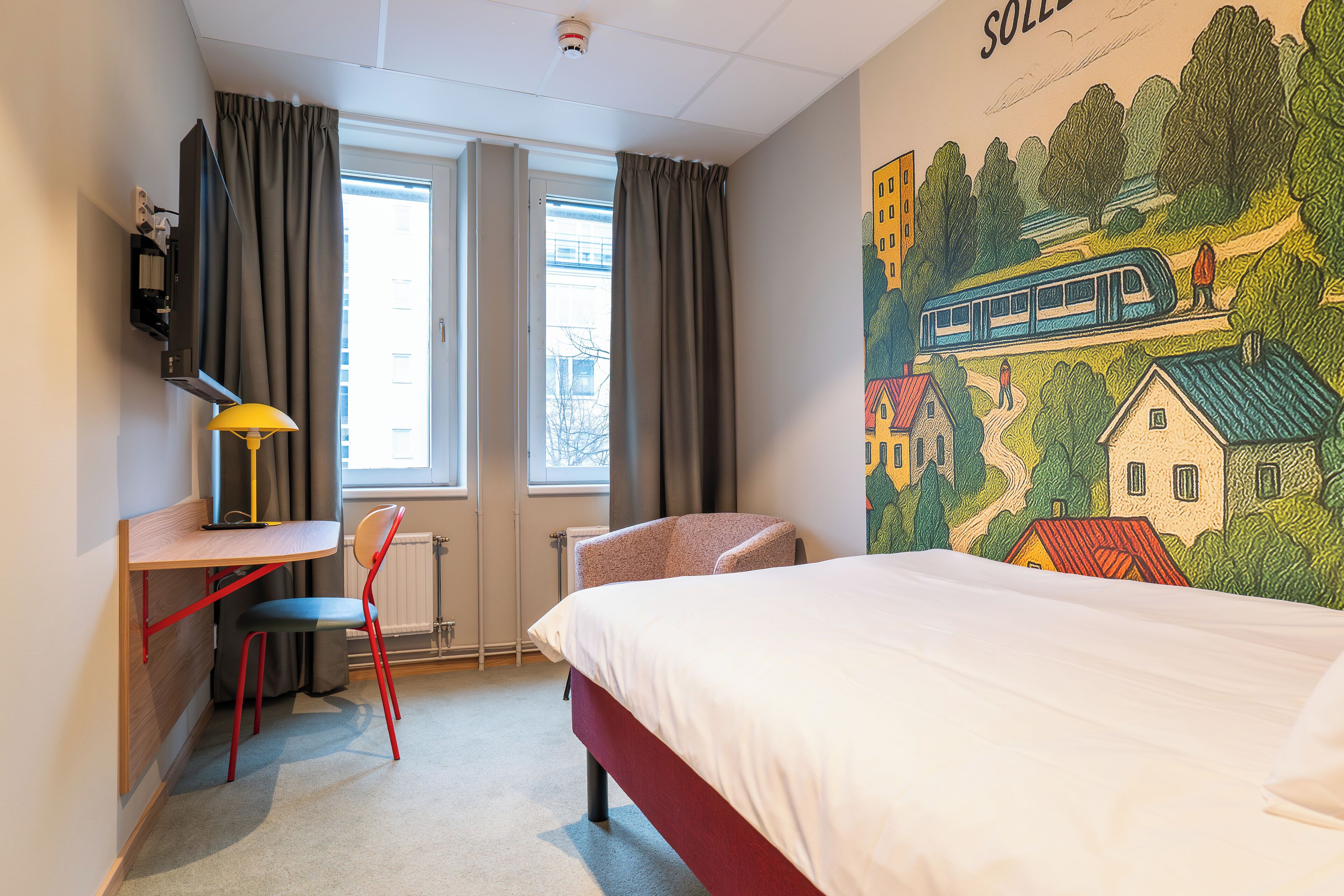 Ein Hotelzimmer mit einem Bett, einem Schreibtisch mit Stuhl, einem Fernseher und einem Fenster mit Vorhängen.