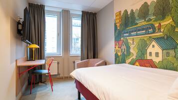 Ein Hotelzimmer mit einem Bett, einem Schreibtisch mit Stuhl, einem Fernseher und einem Fenster mit Vorhängen.