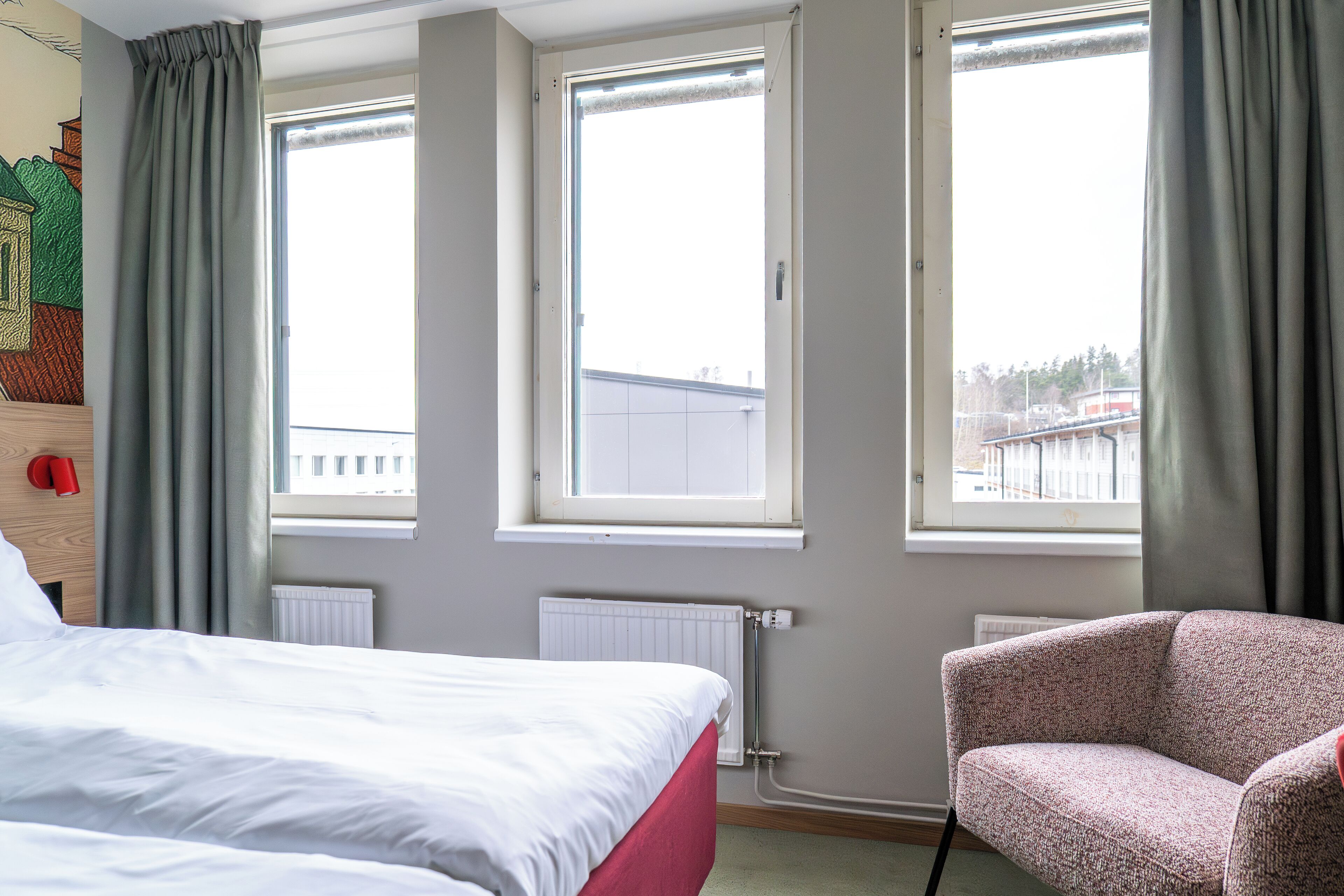 Quarto de hotel com cama, uma cadeira e duas janelas com cortinas.