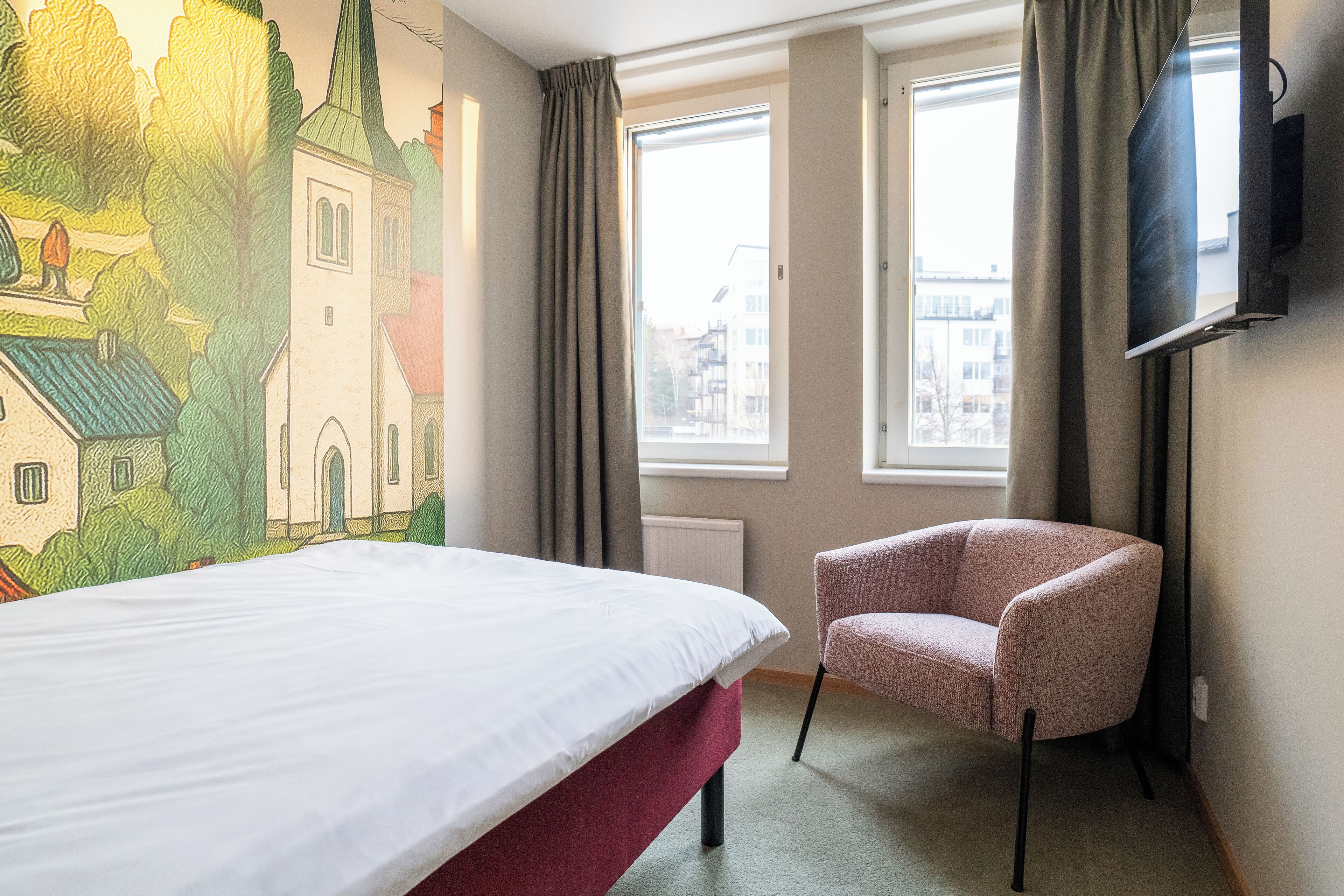 A hotel room with a bed, a chair, a TV, and a mural on the wall.