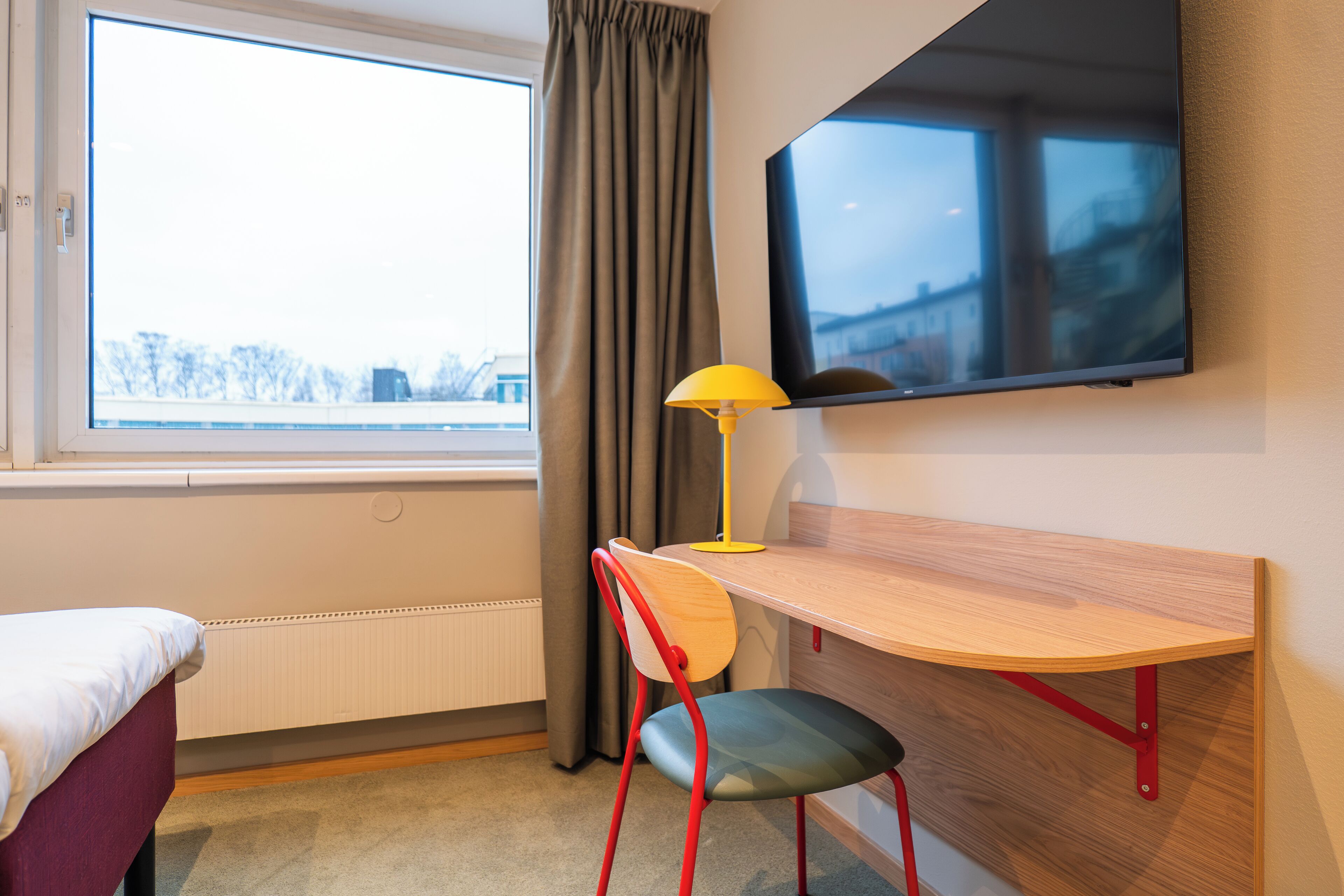 A hotel room with a bed, a desk with a chair, a TV mounted on the wall, and a window with curtains.