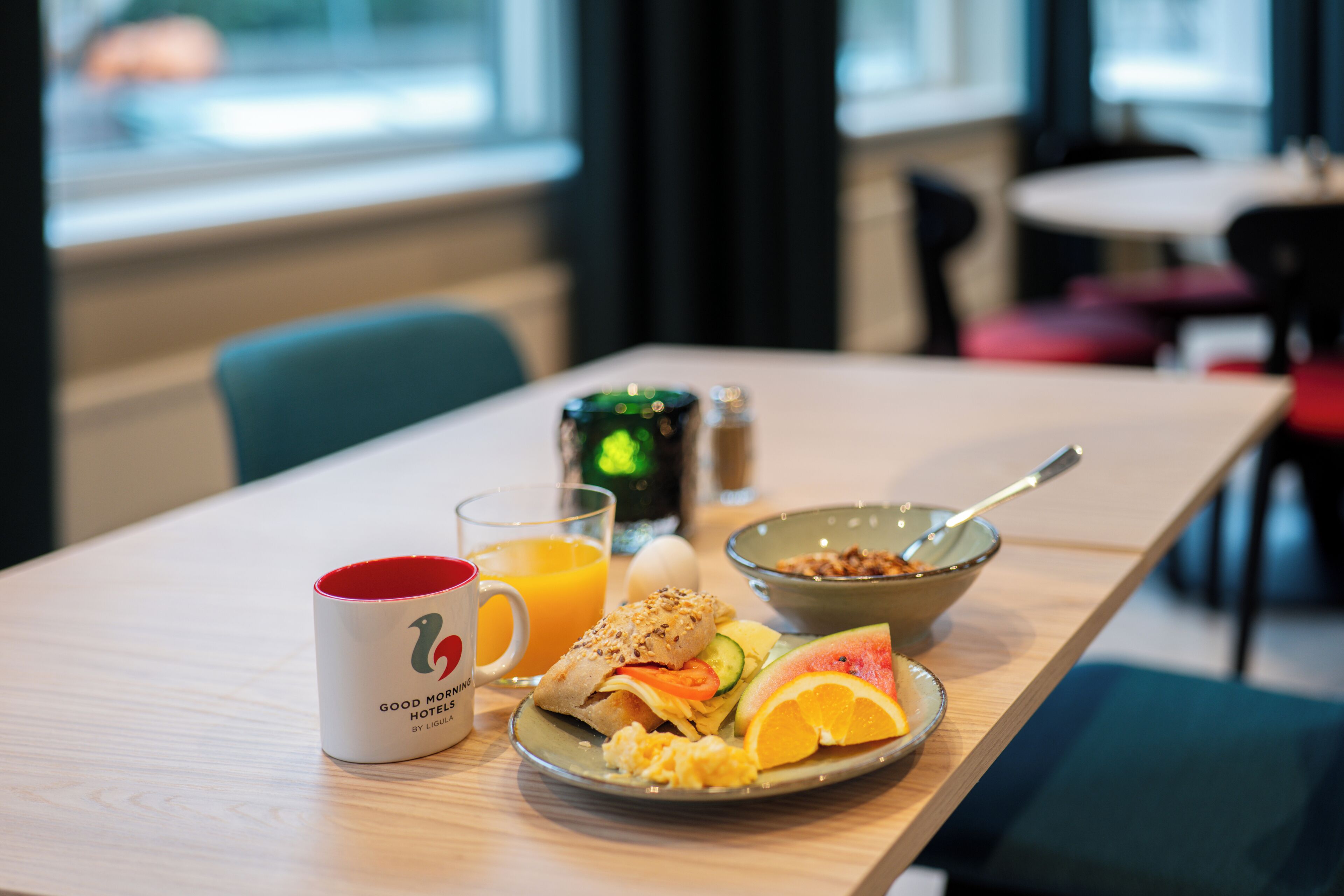 Une table de petit-déjeuner avec une tasse, du jus, un bol de céréales, une assiette d’œufs, des toasts et des fruits.