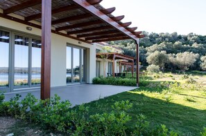 A modern building with large windows, a covered patio area, and a view of a lake. - Thea Houses (South Pelion)