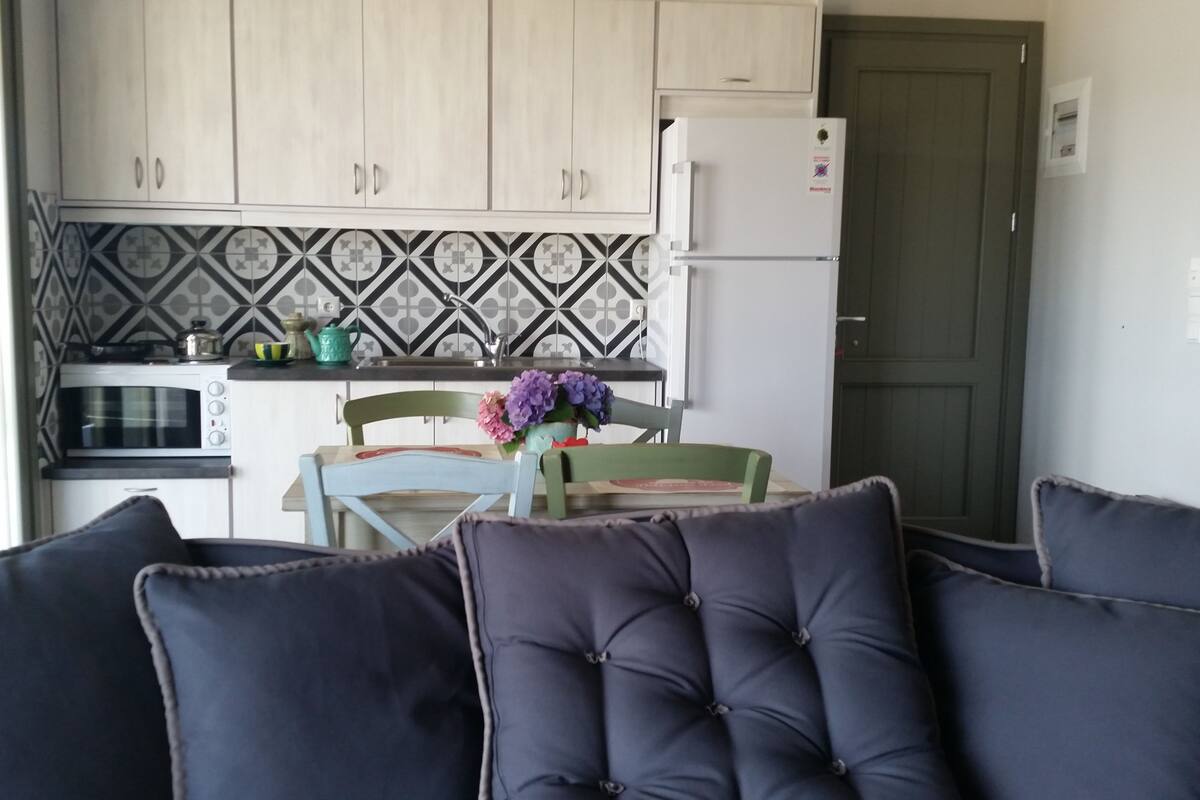 Cocina compacta con microondas, estufa y refrigerador. Hay una mesa de comedor con sillas y un jarrón de flores. La cocina tiene un salpicadero con diseño y gabinetes de color claro.