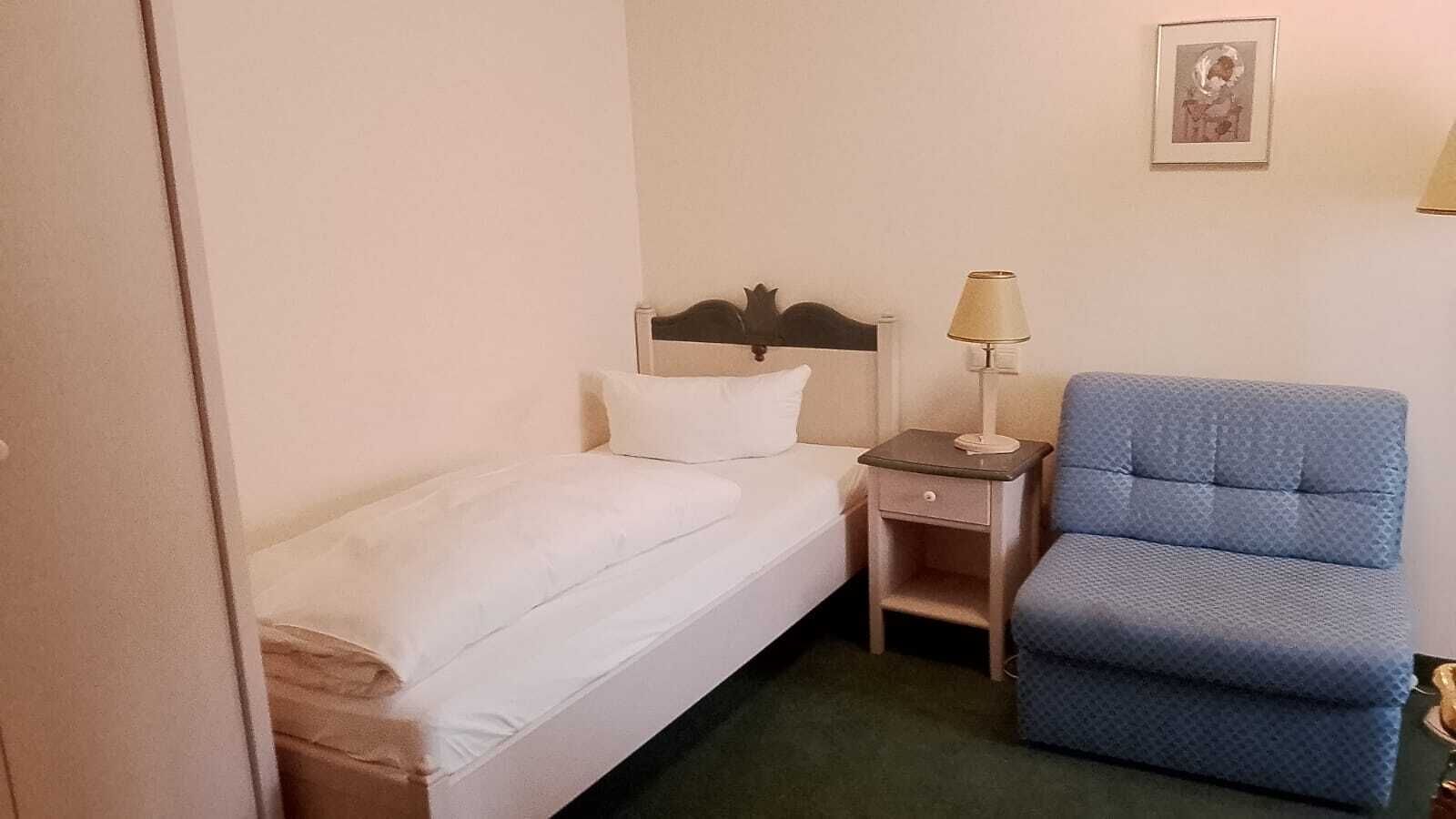 Une petite chambre avec un lit simple, une table de chevet, une lampe et une chaise bleue.
