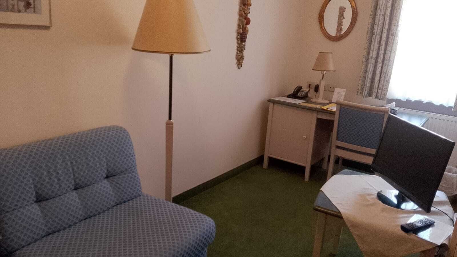 Hotellihuoneessa on sinikuvioinen sohva, työpöytä, jossa on tietokone ja lamppu, pieni kaappi sekä peili seinällä.