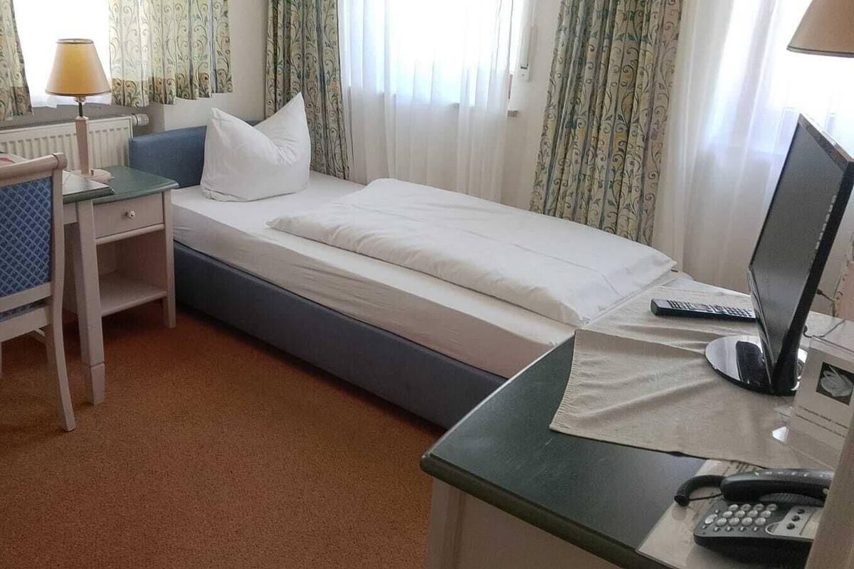 Ein Hotelzimmer mit einem Einzelbett, einem Schreibtisch mit Stuhl, einer Lampe, einem Computer und einem Telefon.