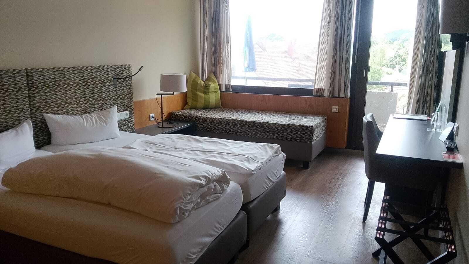 A hotel room with a large bed, a smaller bed, a desk with a chair, and a window with curtains.