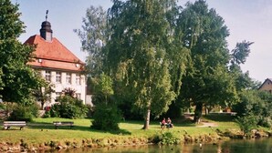 A river with a bridge railing, a large tree, and a building with a red roof. - Hotel-Gasthof Resengörg (Ebermannstadt)