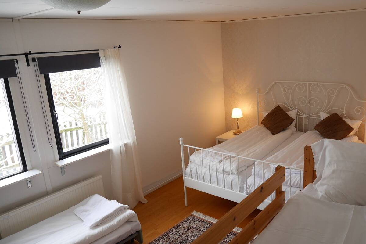 A bedroom with a white metal bed frame, two bedside lamps, a window with curtains, and a small bedside table.