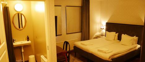 Une chambre d’hôtel avec un lit, une table de chevet avec une lampe, un miroir rond, un lavabo et une chaise.
