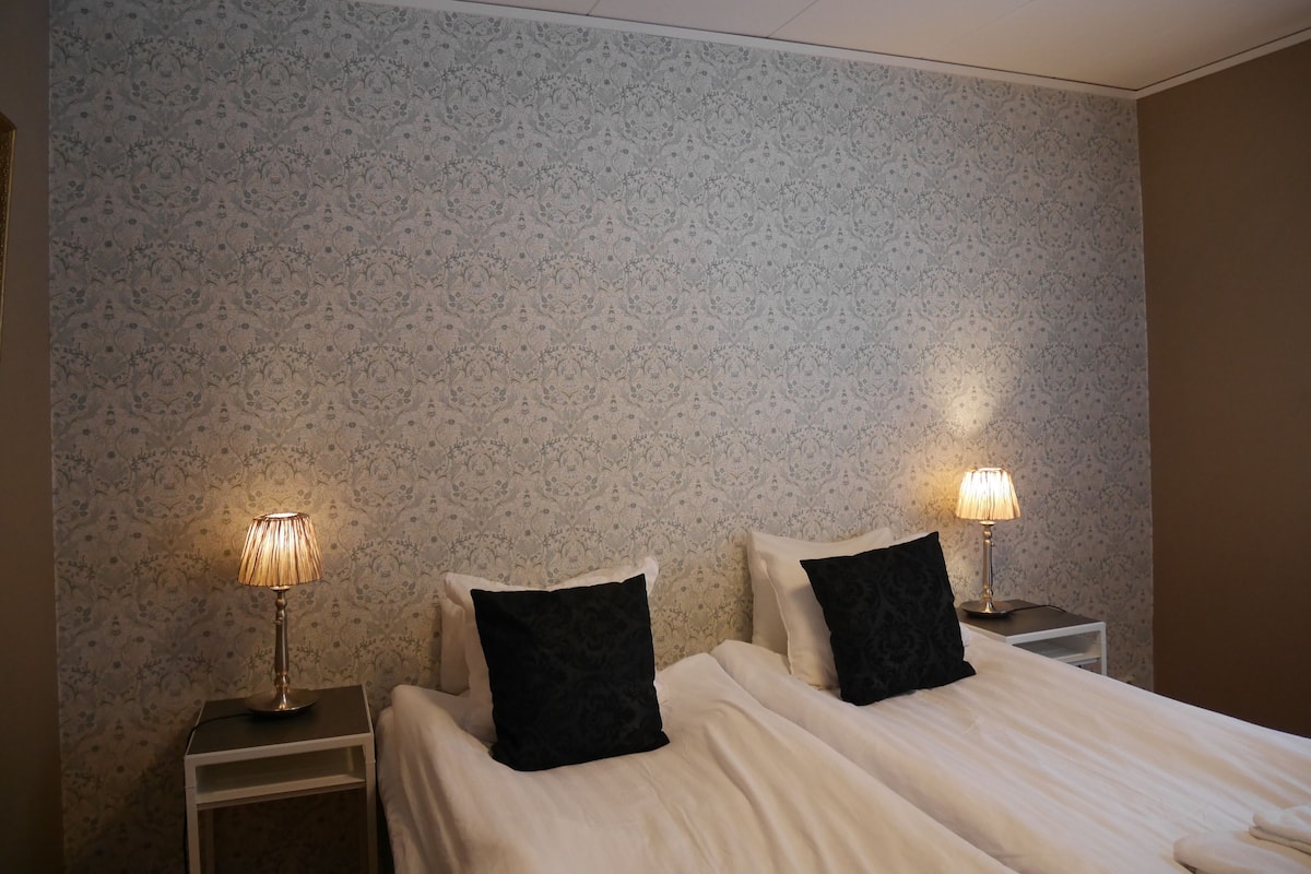 A hotel room with two beds, each with a black pillow and a bedside lamp. The walls have a floral patterned wallpaper.