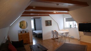 Un cuarto ático acogedor con techo de madera, una mesa de comedor con sillas, un televisor en la pared y una cama en la habitación contigua.