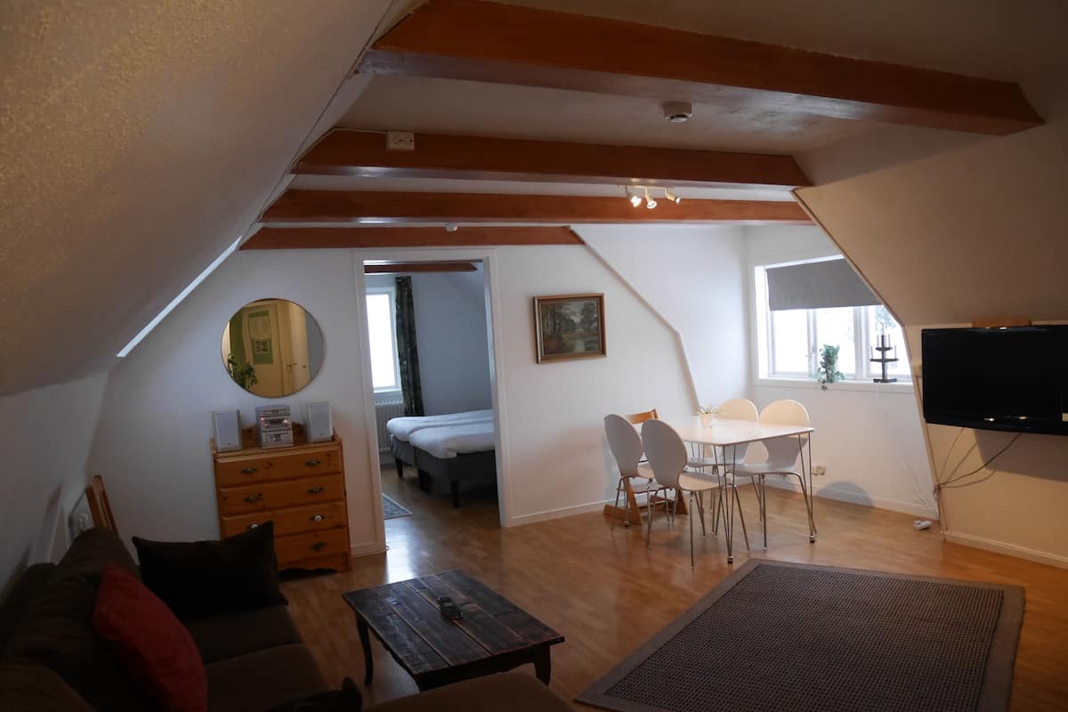 A cozy attic room with a wooden ceiling, a dining table with chairs, a TV mounted on the wall, and a bed in the adjacent room.