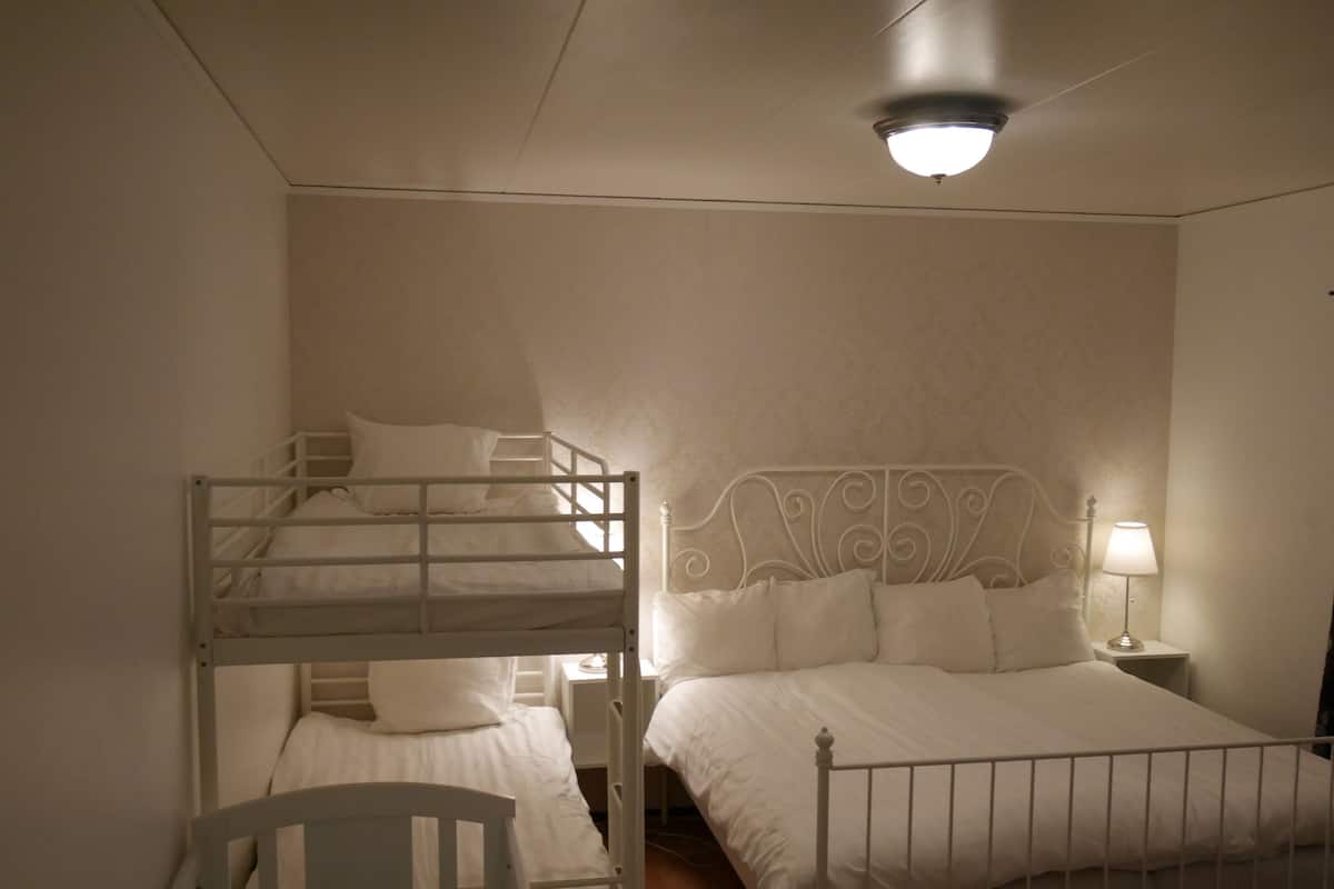 A bedroom with a bunk bed and a single bed, both with white bedding. There is a nightstand with a lamp and a ceiling light.