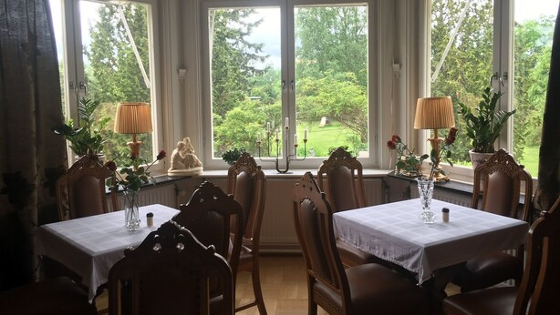 Comedor con ventanal que da a un jardín. Mesas con manteles blancos y sillas de madera alrededor.
