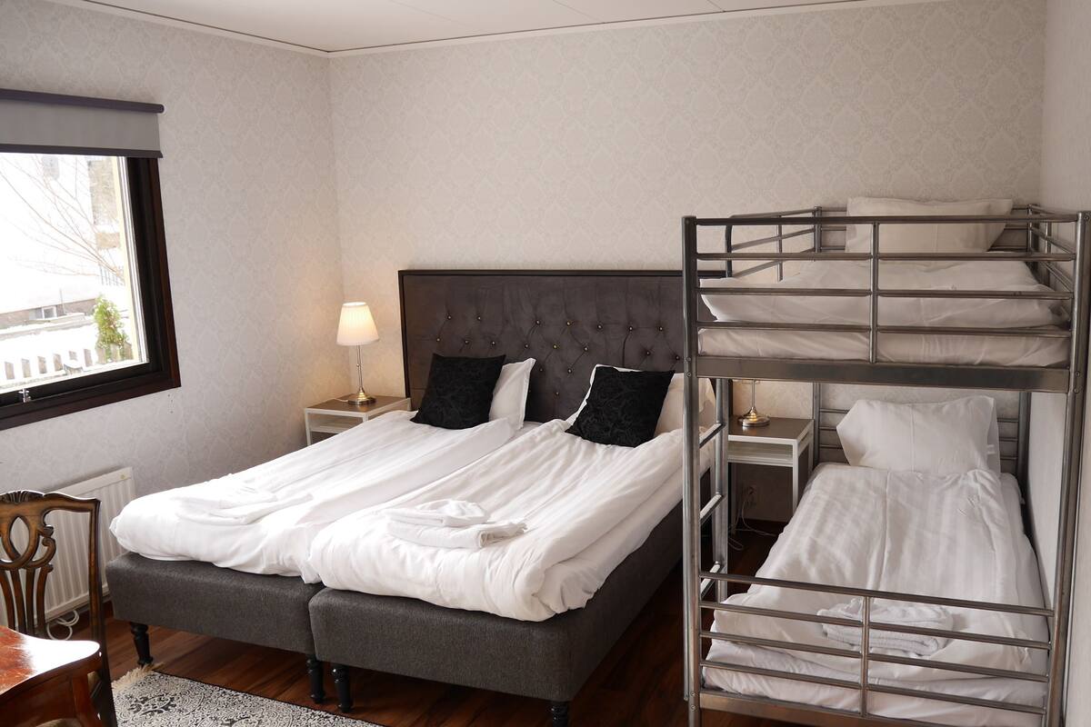 A hotel room with a single bed, a bunk bed, a nightstand with a lamp, a wooden chair, and a window with a view.