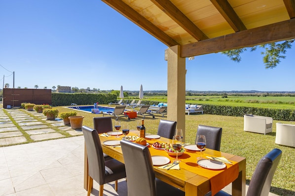 A covered outdoor dining area with a pool and lounge chairs in the background.