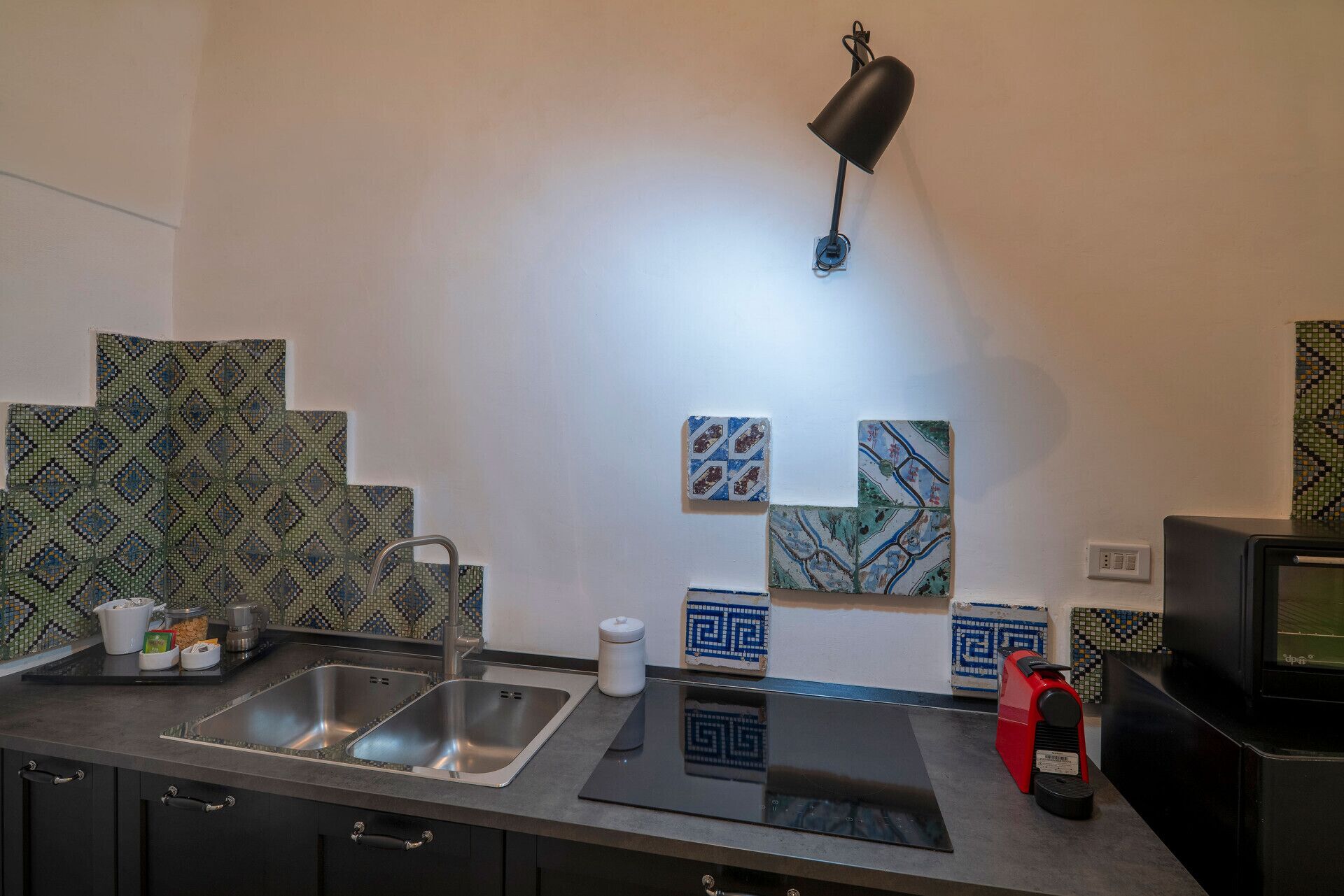 Una cocina con un lavamanos, una cafetera y un microondas. Hay azulejos decorativos en la pared y un televisor pequeño sobre la encimera.