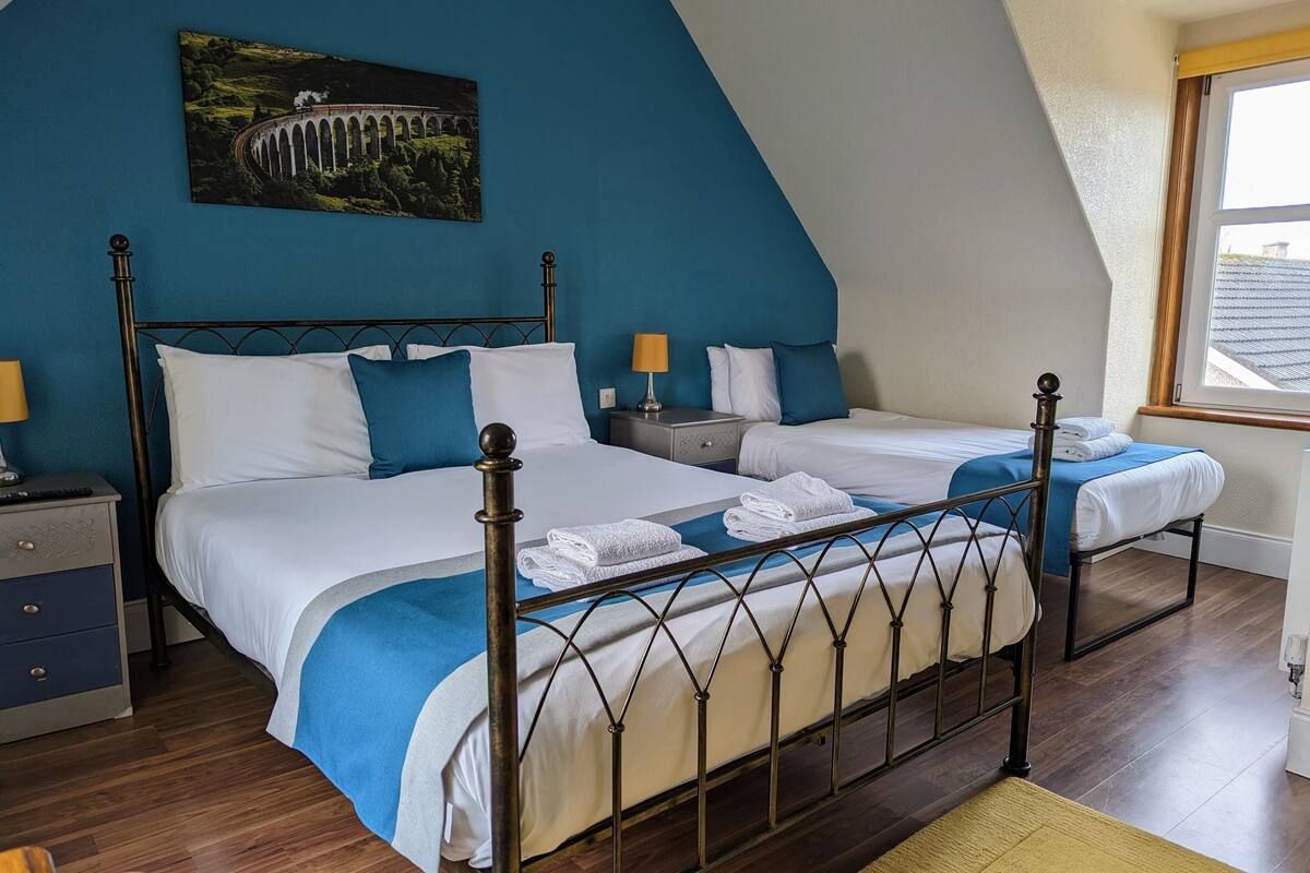 A bedroom with two beds, a nightstand, a lamp, and a painting of a bridge.