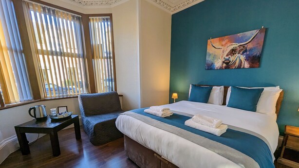 A hotel room with a large bed, a chair, a small table with a kettle, and a painting on the wall.