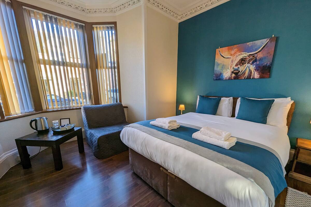 A hotel room with a large bed, a chair, a small table with a kettle, and a painting on the wall.