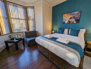A hotel room with a large bed, a chair, a small table with a kettle, and a painting on the wall. - The Coo's Guest House (Inverness)