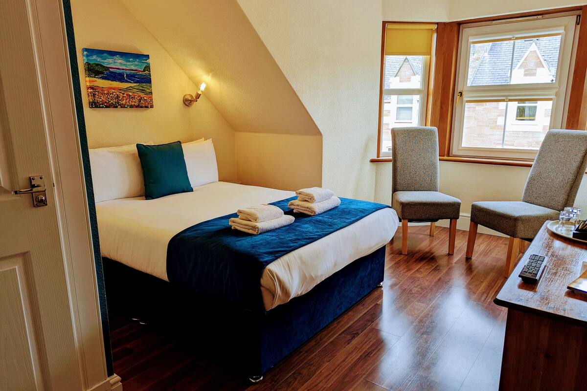 A bedroom with a bed, two chairs, a desk, a painting, and a window.