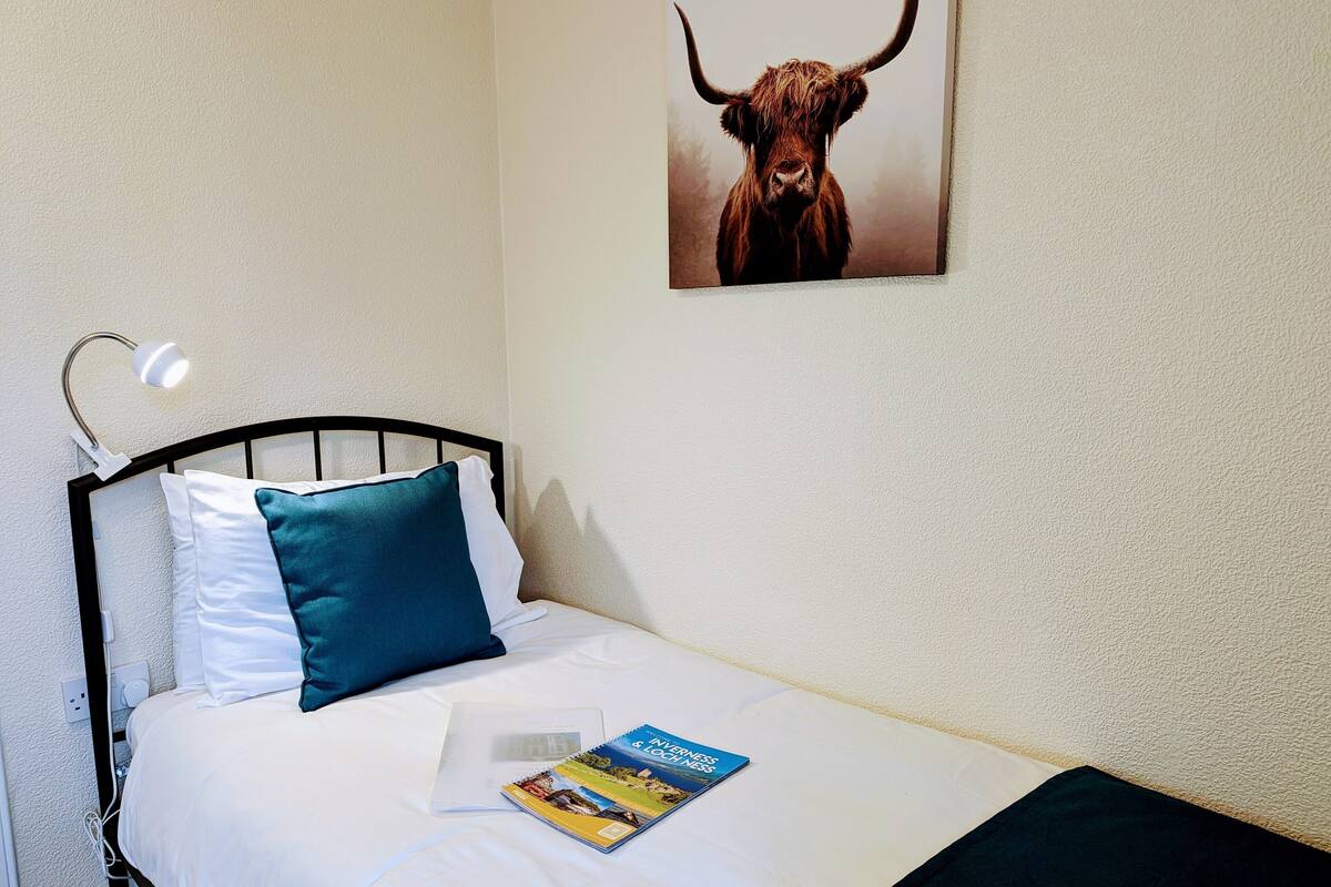 A neatly made bed with white and teal pillows, a book titled "Innersness with Ness," and a framed picture of a Highland cow on the wall.