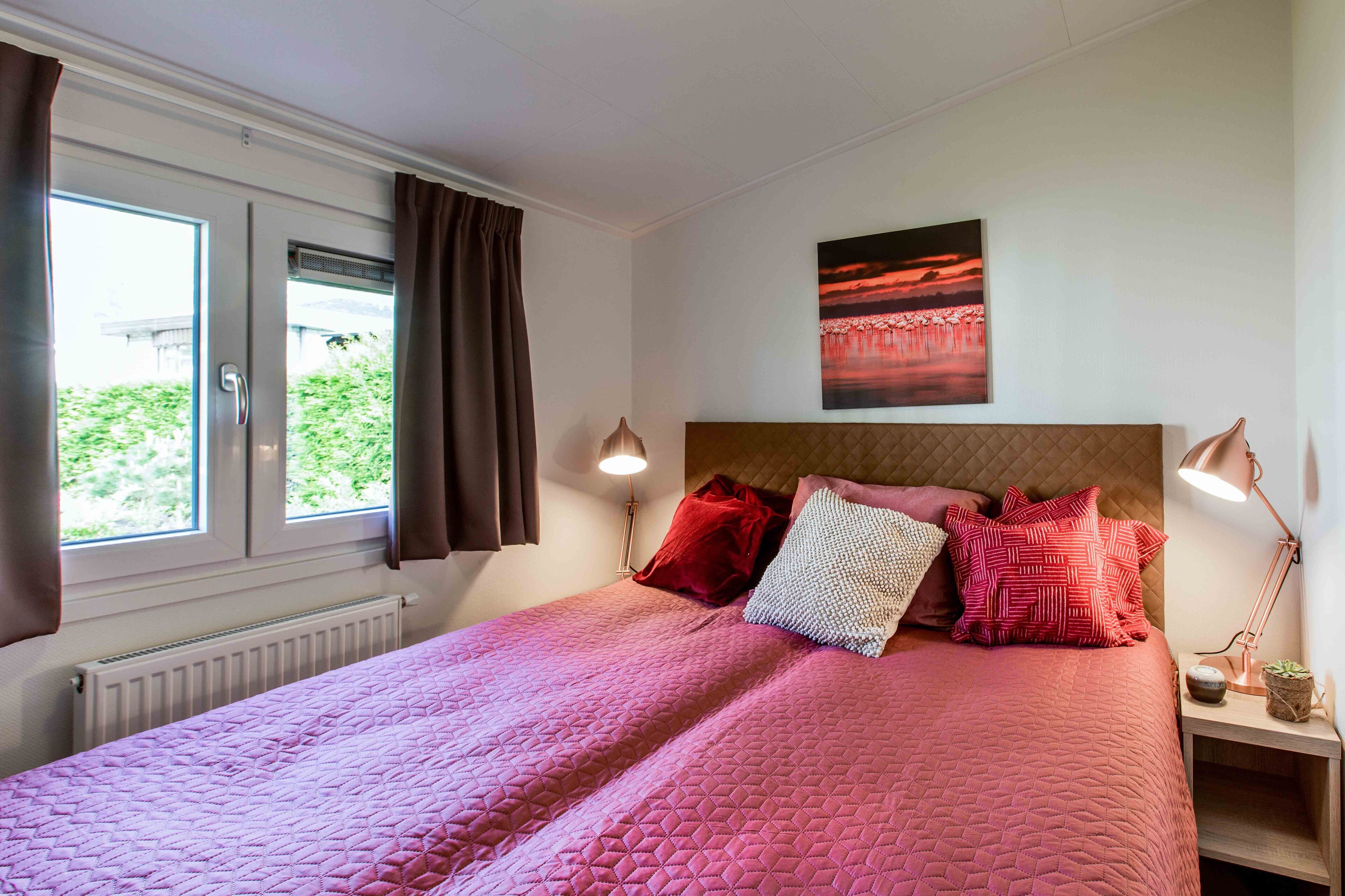 A neatly made bed with a pink quilt, a red pillow, and a white pillow. There is a painting of a sunset on the wall, a window with curtains, and a bedside lamp.