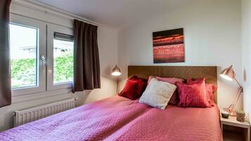 A neatly made bed with a pink quilt, a red pillow, and a white pillow. There is a painting of a sunset on the wall, a window with curtains, and a bedside lamp.
