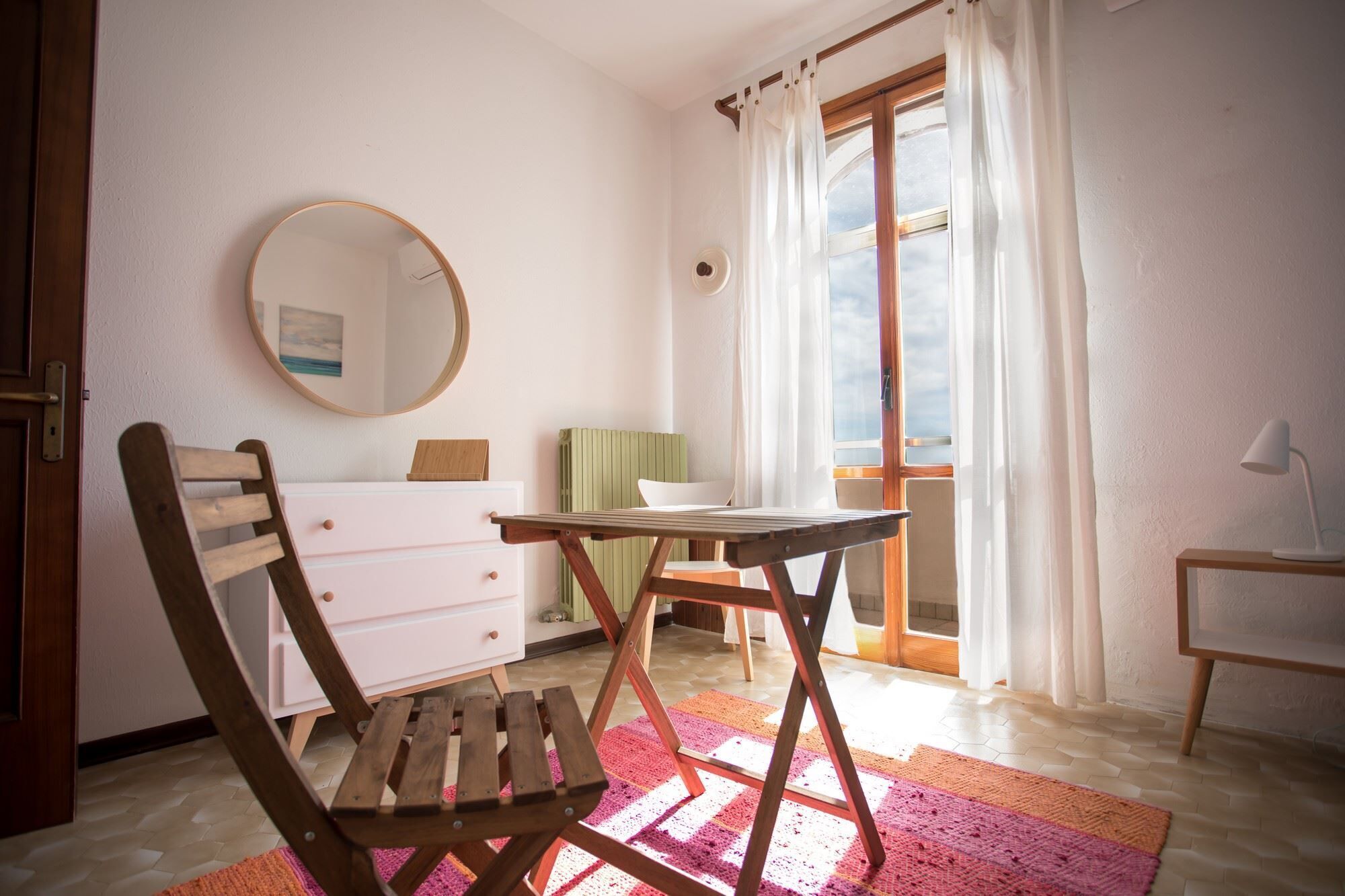 Un dormitorio con una silla de madera, una mesita pequeña, un aparador, un espejo y una ventana con cortinas.