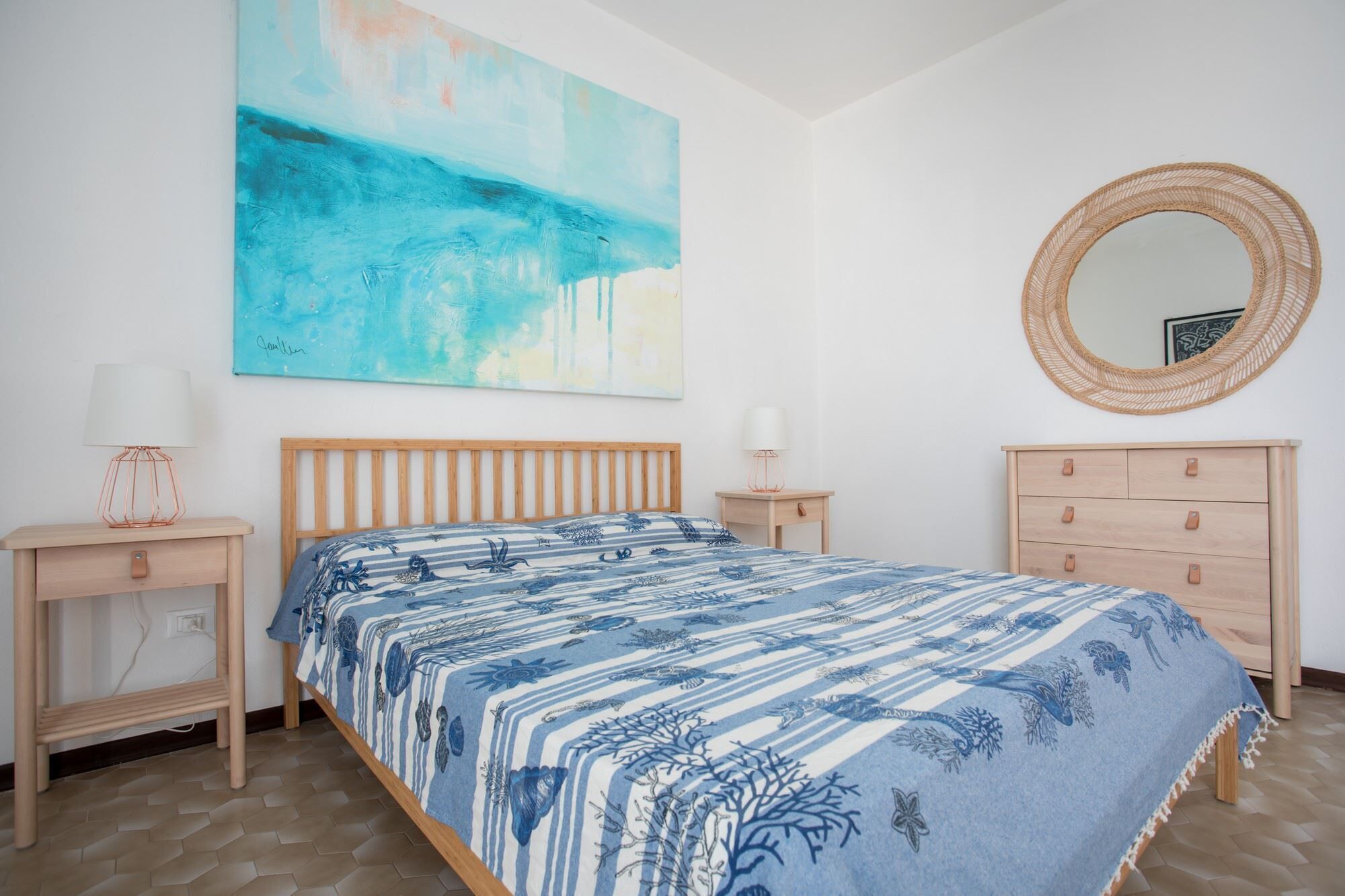 Un dormitorio con cama, mesita de noche, cómoda, espejo redondo y un cuadro abstracto grande en la pared.