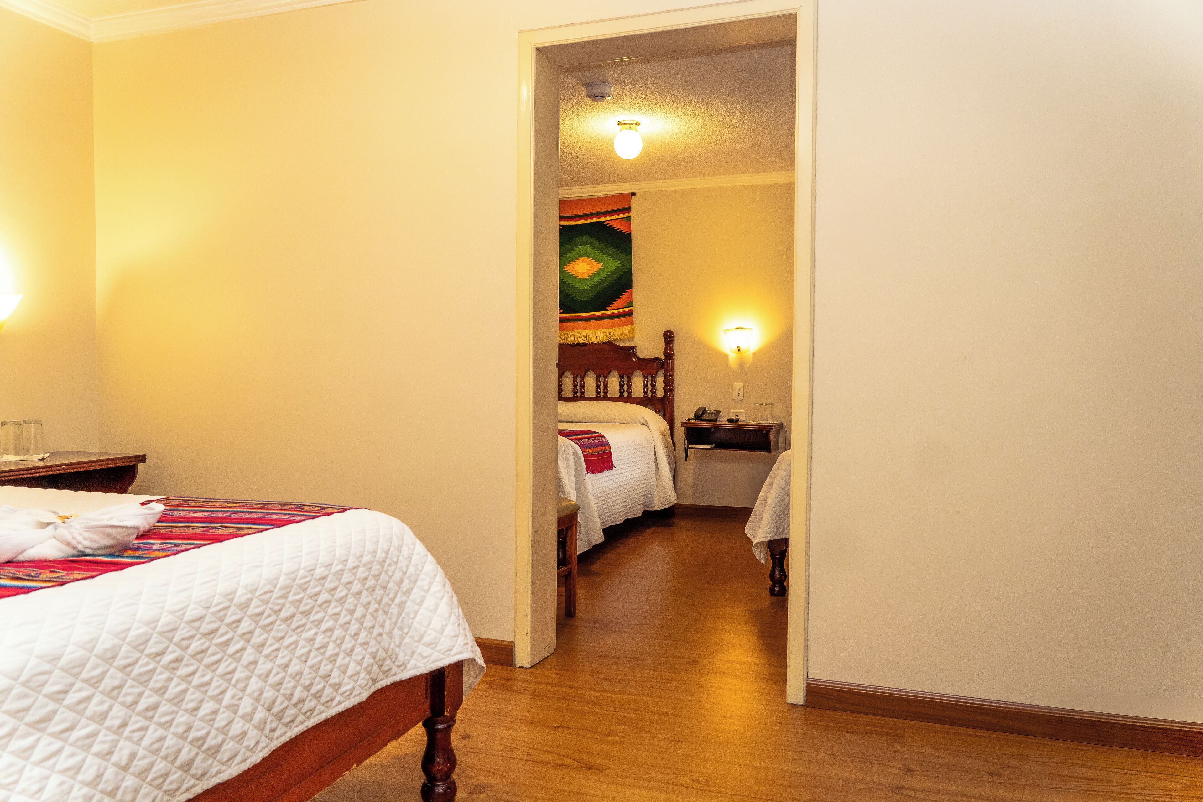 Una habitación de hotel con una cama de madera, una mesita de noche y otra mesilla auxiliar. Se ve una puerta que da a otra habitación.