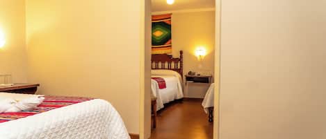 Una habitación de hotel con una cama de madera, una mesita de noche y otra mesilla auxiliar. Se ve una puerta que da a otra habitación.