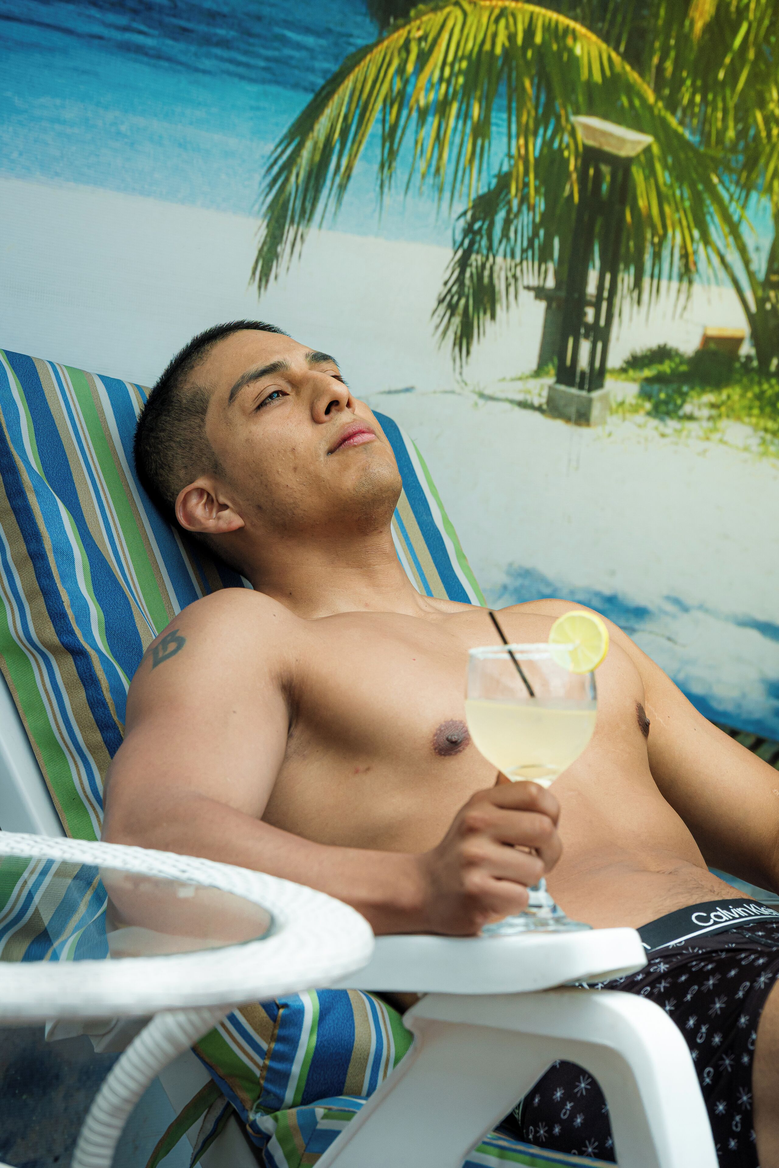 A person lying on a striped beach-themed lounge chair, holding a cocktail with a lemon slice, with a palm tree and ocean in the background.