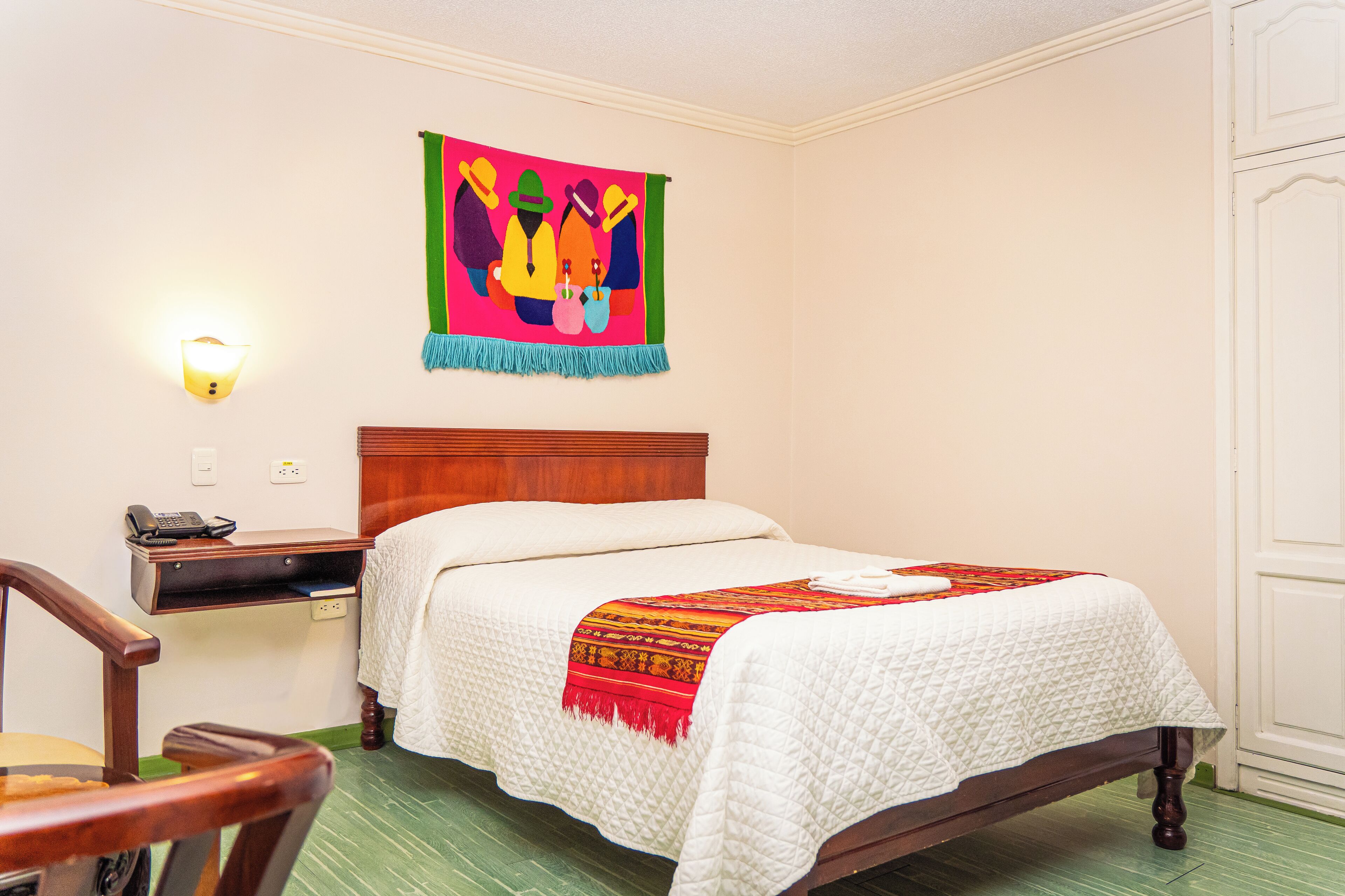 A hotel room with a wooden bed, a bedside table with a lamp, a chair, and a colorful tapestry on the wall.