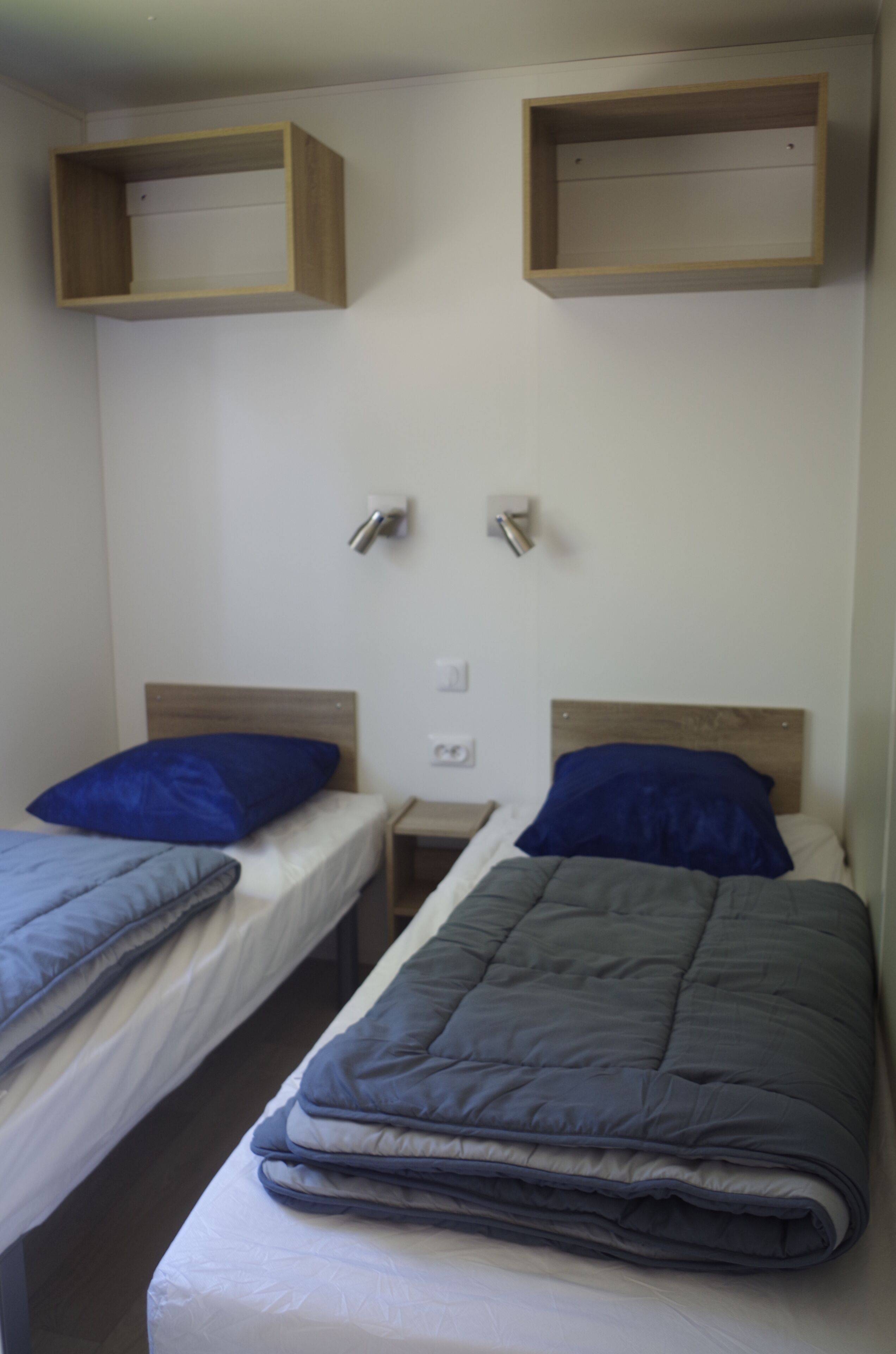A room with two single beds, each with a wooden headboard and a blue pillow. There are two wall-mounted light switches and a small wooden shelf above the beds.