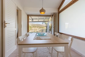 A dining area with a wooden table and white chairs, a hanging light fixture, and a view of a hillside through large windows.