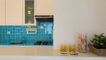 Une cuisine avec un plan de travail recouvert de carreaux bleus, une cuisinière à gaz, un évier et une fenêtre. On y trouve deux verres de jus d’orange et un panier de pain sur le comptoir.