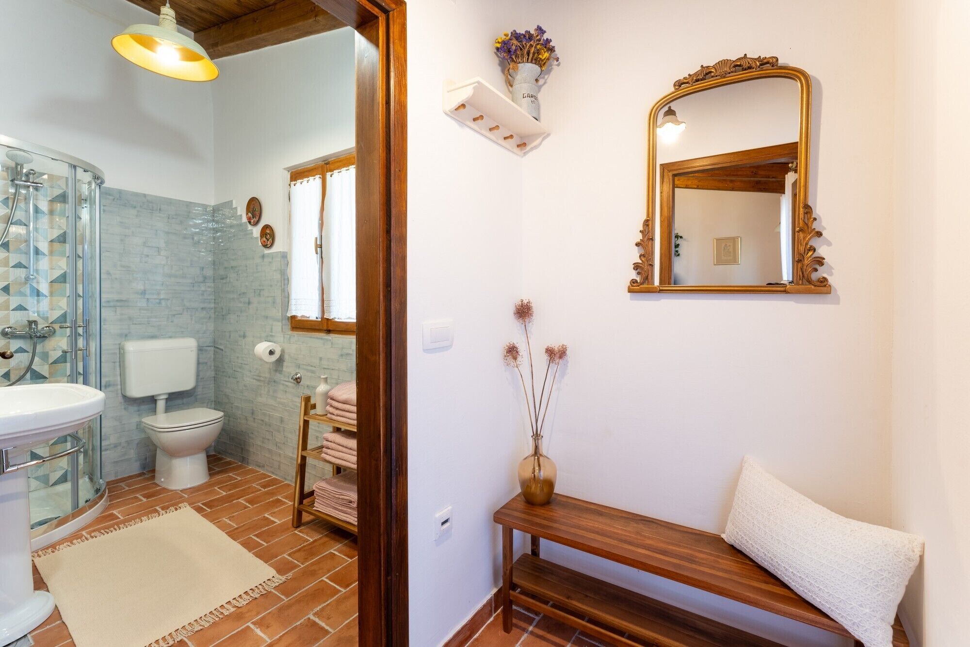 A bathroom with a glass shower enclosure, a white sink, and a toilet. There is a wooden bench with a white cushion and a vase with dried flowers on the wall.