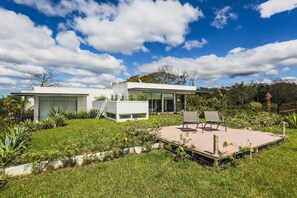 Une maison moderne dotée d’un patio, entourée d’une végétation luxuriante et surplombant un ciel d’un bleu éclatant.