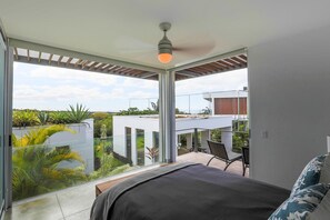 Une chambre à coucher moderne, dotée d’un grand lit, d’un ventilateur de plafond et offrant une vue sur un jardin et les bâtiments voisins.