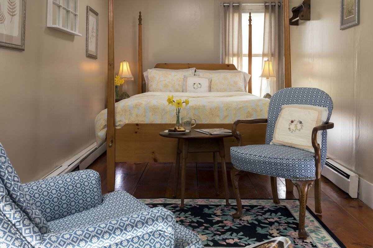 A bedroom with a four-poster bed, a wooden chair, a small table with a vase of flowers, and a floral rug.