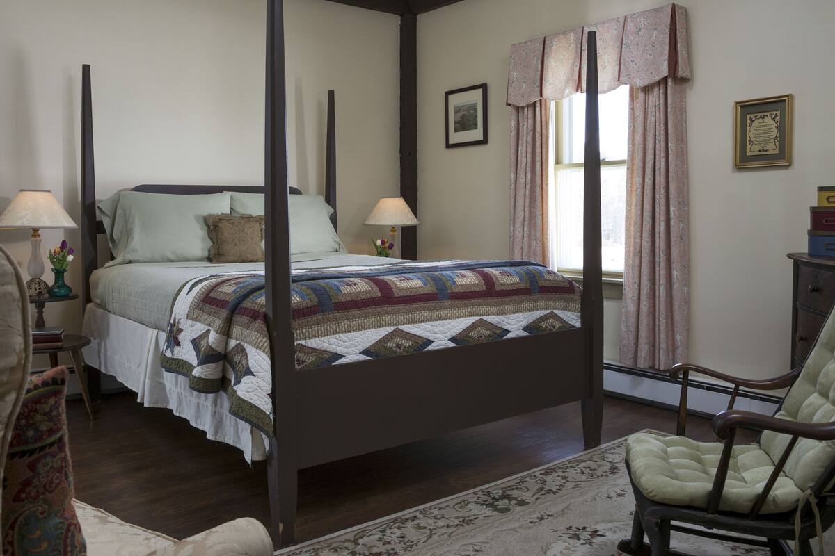 A four-poster bed with a quilt, a chair, and a nightstand with a lamp.