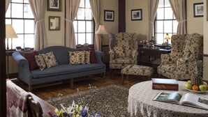 Una sala de estar acogedora con un sofá azul, sillones estampados, una mesa de centro de madera con un libro y fruta, y un jarrón floral.